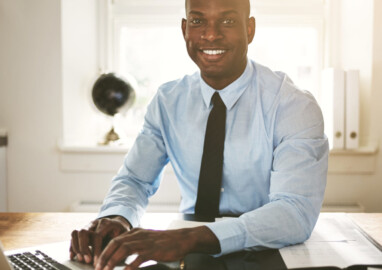 Successful young African businessman sitting at his desk in an office smiling and working on a laptop
