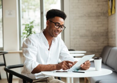 Picture of happy young african man sitting coworking with tablet computer. Looking aside.