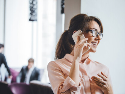 Woman having a conversation on a smartphone. There is a man in the background who is wearing a suit. He is being served tea by a waiter.