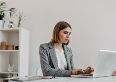 Attractive blonde woman typing letter in MacBook at her workplace. Photo of lady in stylish jacket in bright office