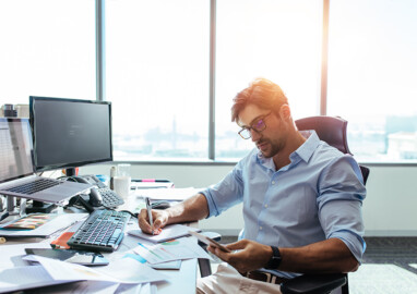 Entrepreneur making notes while looking at mobile phone. Young businessman sitting at his desk in office working with papers while seated in front of desktop computers.