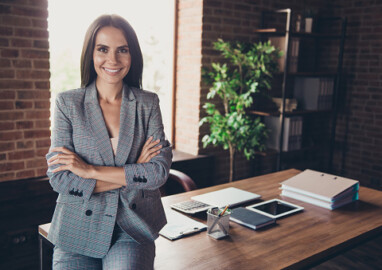 Beautiful elegant classic trendy latin smiling lady freelancer, company founder with folded crossed hands, wearing gray suit, invites to her team, at work place, station, modern fashionable loft