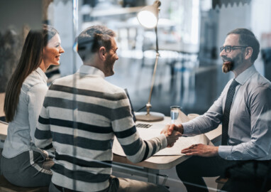 Happy real estate agent shaking hands with young couple after successful meeting in the office. The view is through glass.